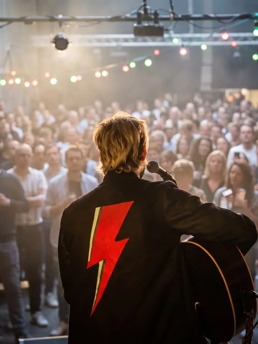 Juri Volta von hinten auf der Bühne, Blitz auf der Jacke, vor vollem Saal